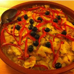 Caçarola de bacalhau assada numa travessa de barro, coberta com tiras de pimento vermelho assado e azeitonas pretas