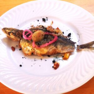 Peixe frito inteiro coberto com rodelas de cebola roxa em conserva, azeitonas e crocante preto num prato branco