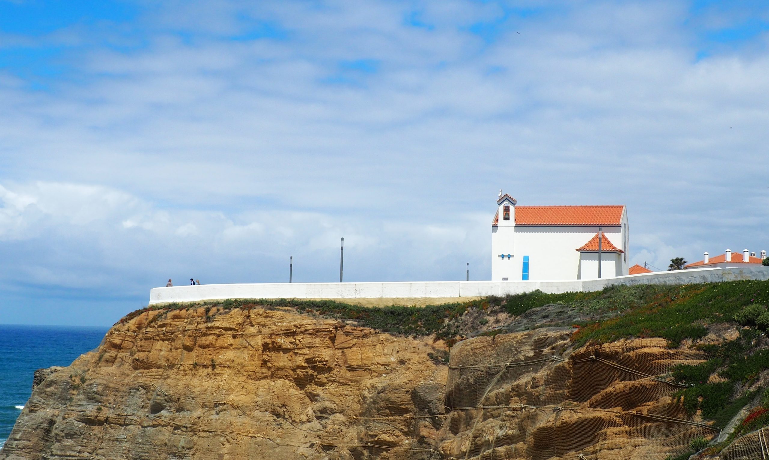 Pequeña capilla blanca con tejado de tejas naranjas encaramada a un acantilado costero rocoso con vistas al océano Atlántico.