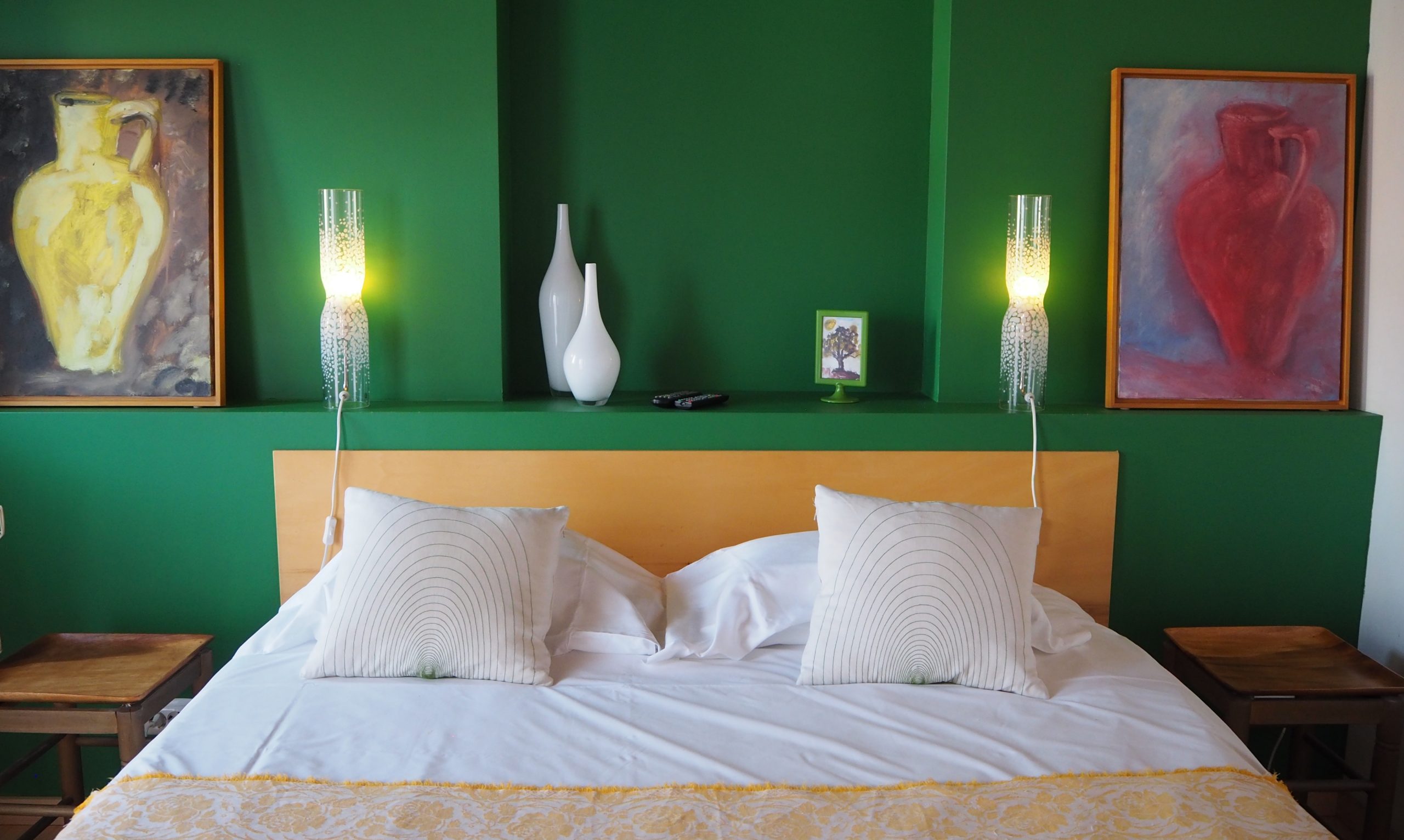 Habitación de hotel muy colorida con una cama de matrimonio, una pared decorativa en verde, dos cuadros de jarrones y objetos decorativos de cerámica blanca en una estantería