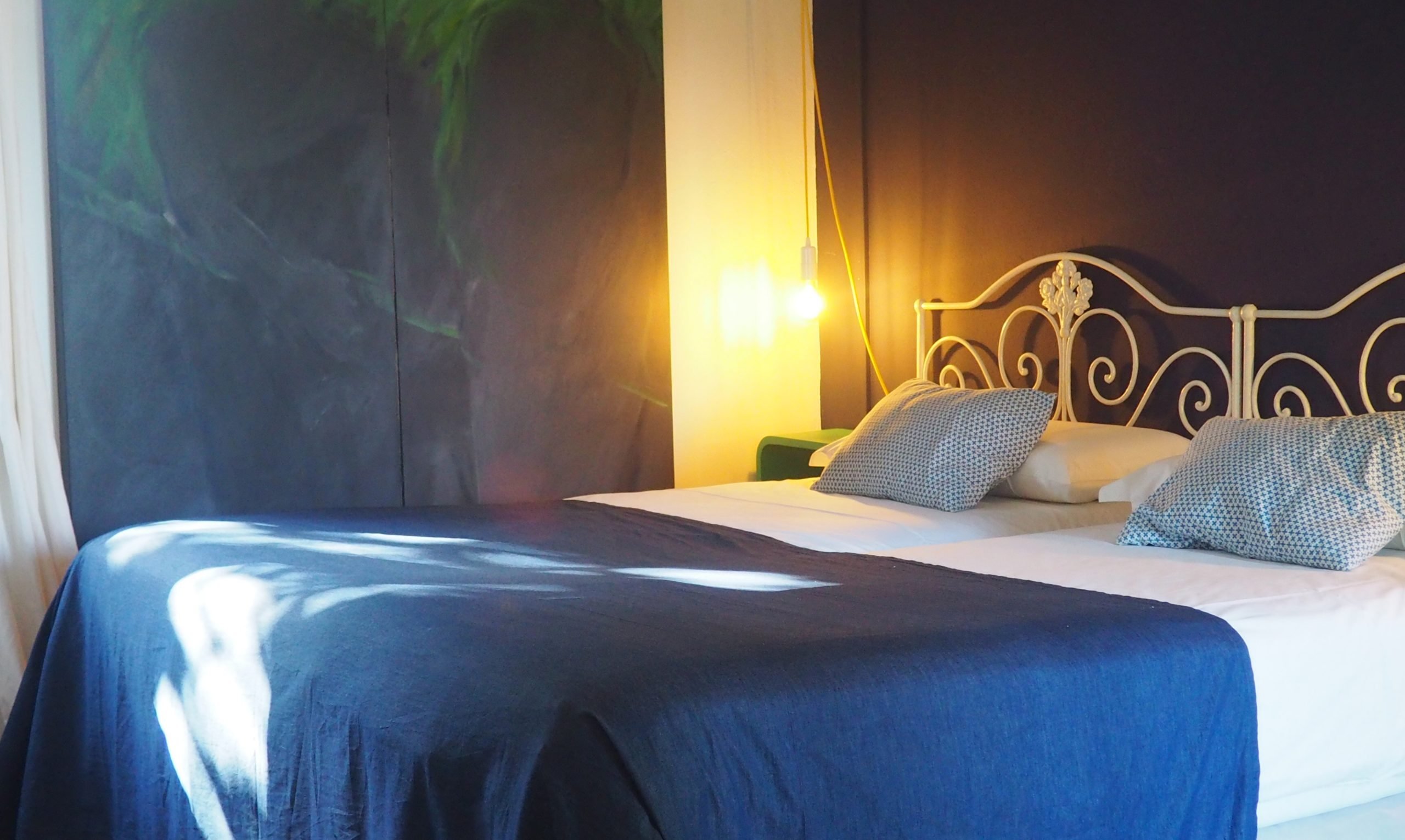 Dormitorio con una cama de matrimonio, cabecero de hierro forjado blanco con adornos, ropa de cama azul y una lámpara colgante con bombilla Edison sobre una pared oscura
