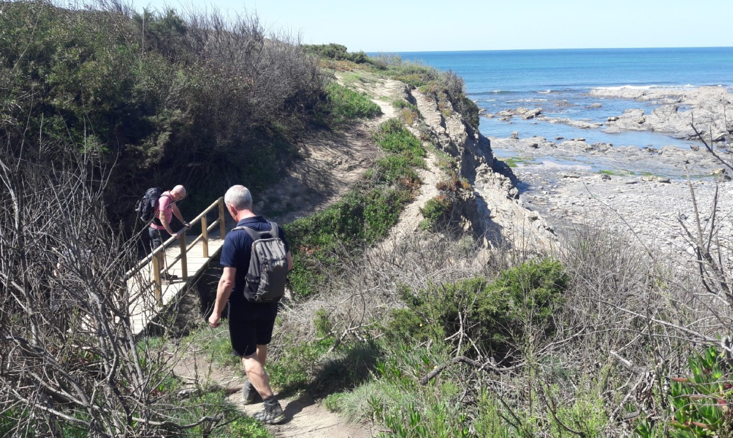 Dos excursionistas con mochilas cruzando un pequeño puente de madera en un sendero costero que discurre por encima de la costa rocosa y el mar azul