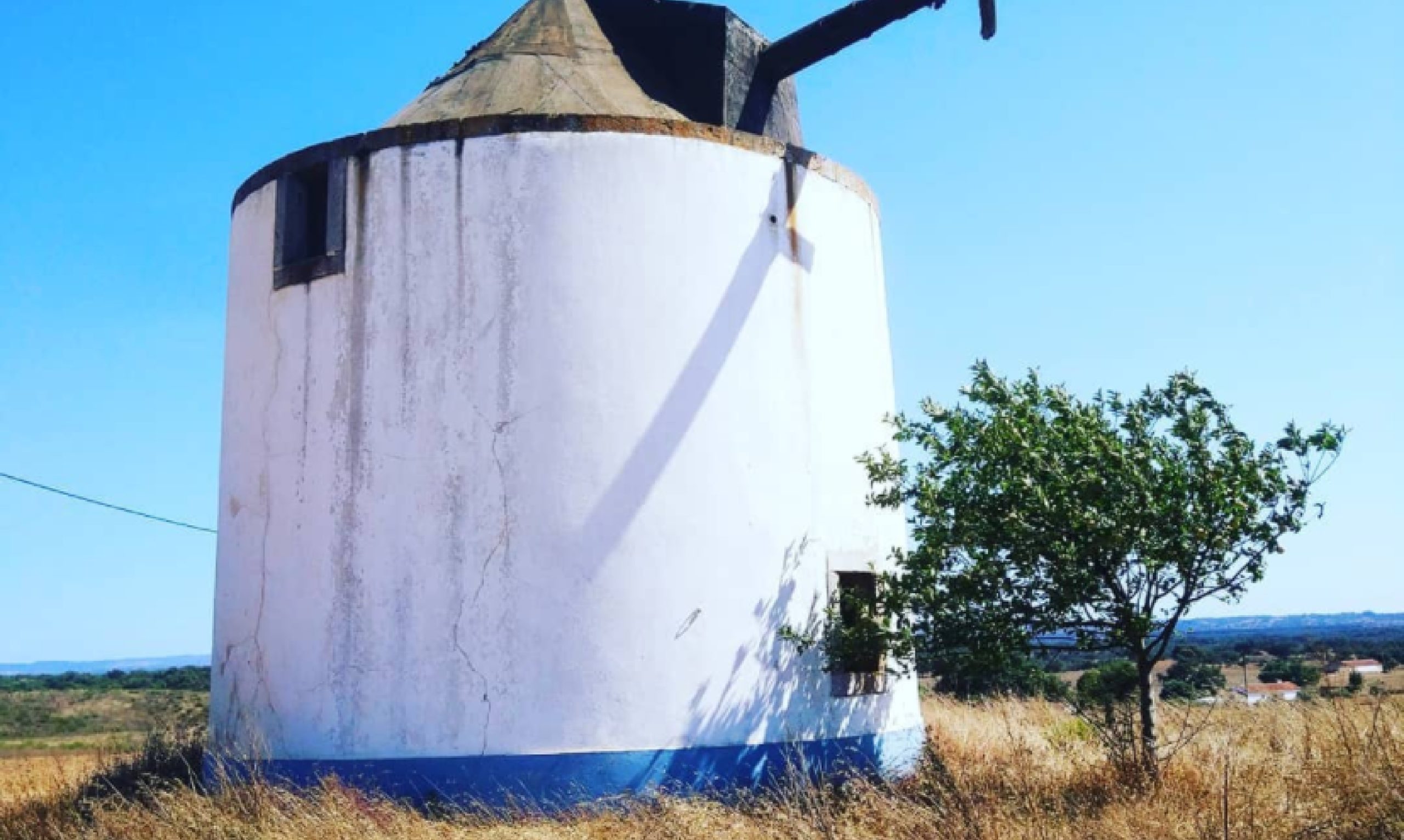 Antigua torre de molino de viento blanca con tejado cónico y un ala parcialmente conservada, situada en un paisaje rural árido bajo un cielo azul y despejado