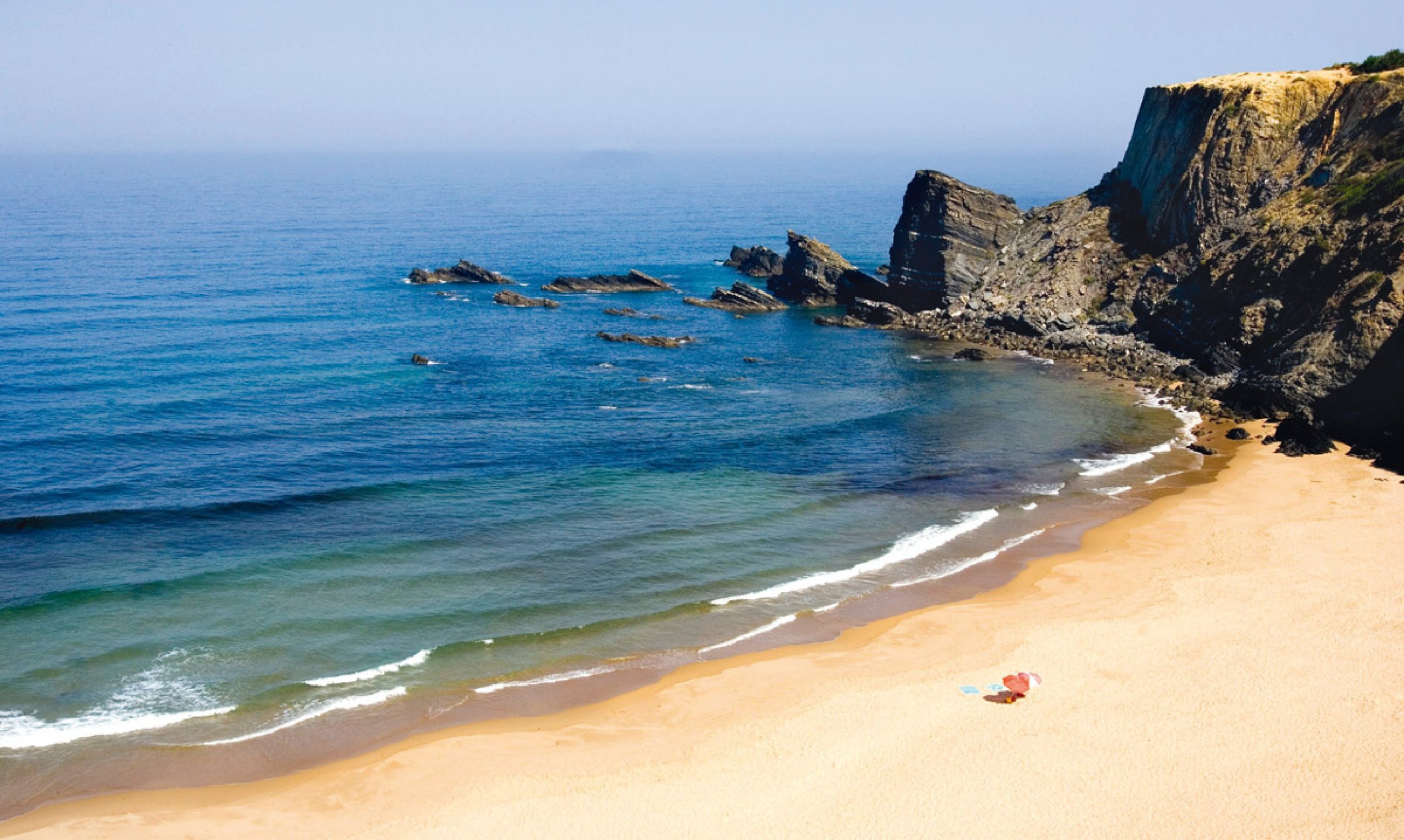 Una playa de arena apartada con acantilados rocosos, rocas dispersas en el mar y tranquilas olas azules