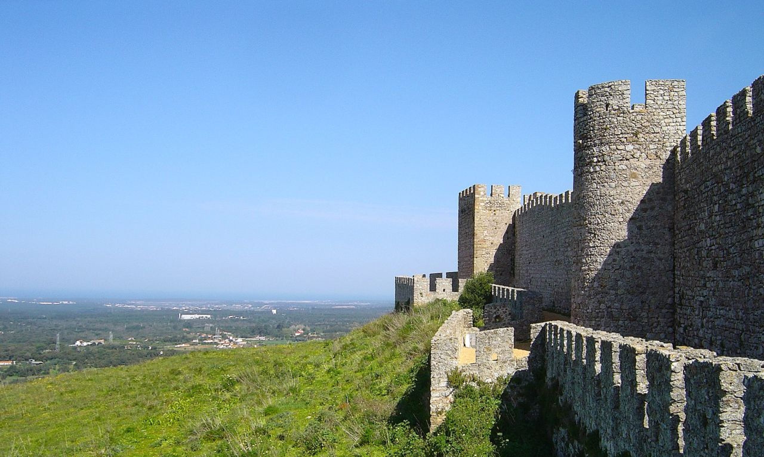 Muros de piedra de un castillo medieval y torres almenadas en lo alto de una colina, con vistas a un valle verde y a la costa en la lejanía, bajo un cielo azul y despejado