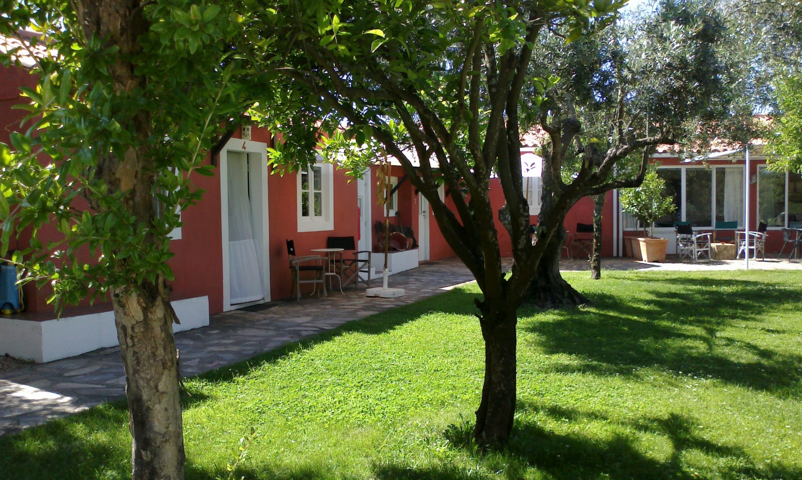 Exterior de un edificio rojo con puertas con marcos blancos, un patio con baldosas y asientos, y un exuberante césped verde con árboles