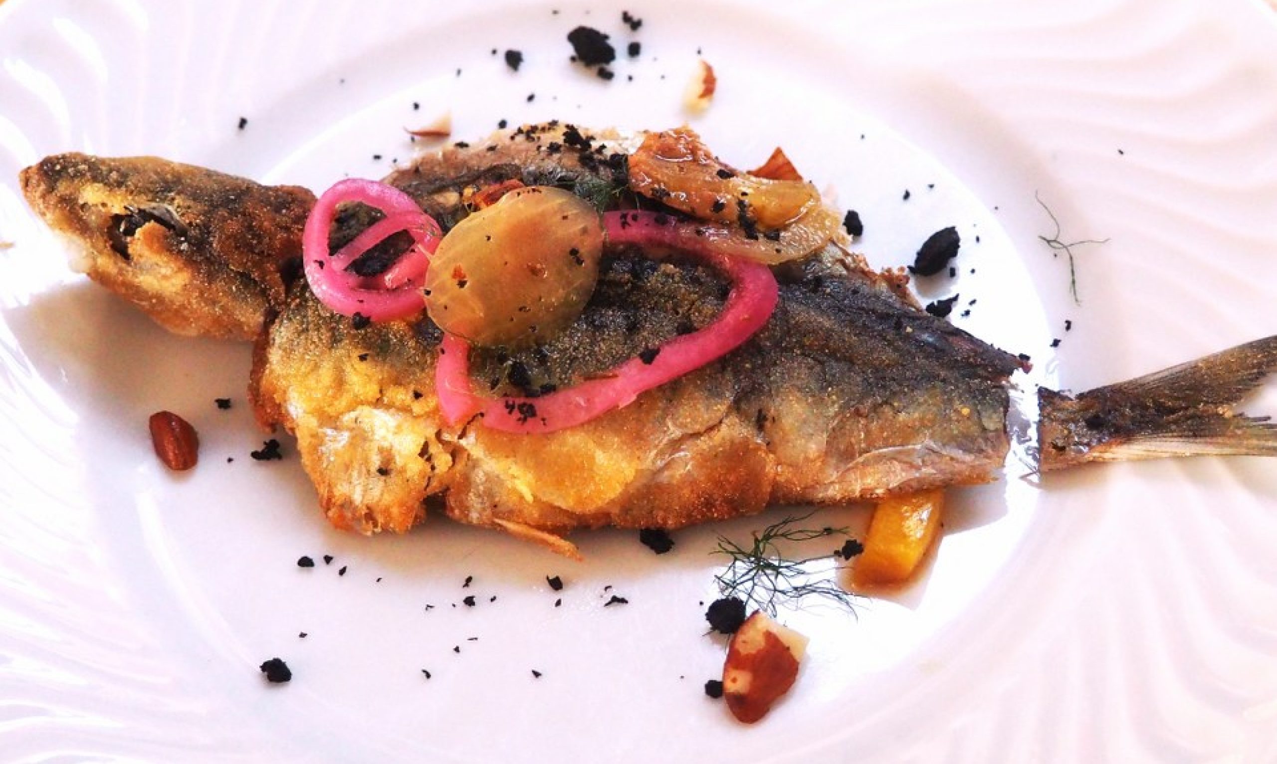 Pescado frito entero cubierto con cebolla roja encurtida, aceitunas y cebolla caramelizada, adornado con eneldo y migas de pan negro en un plato blanco
