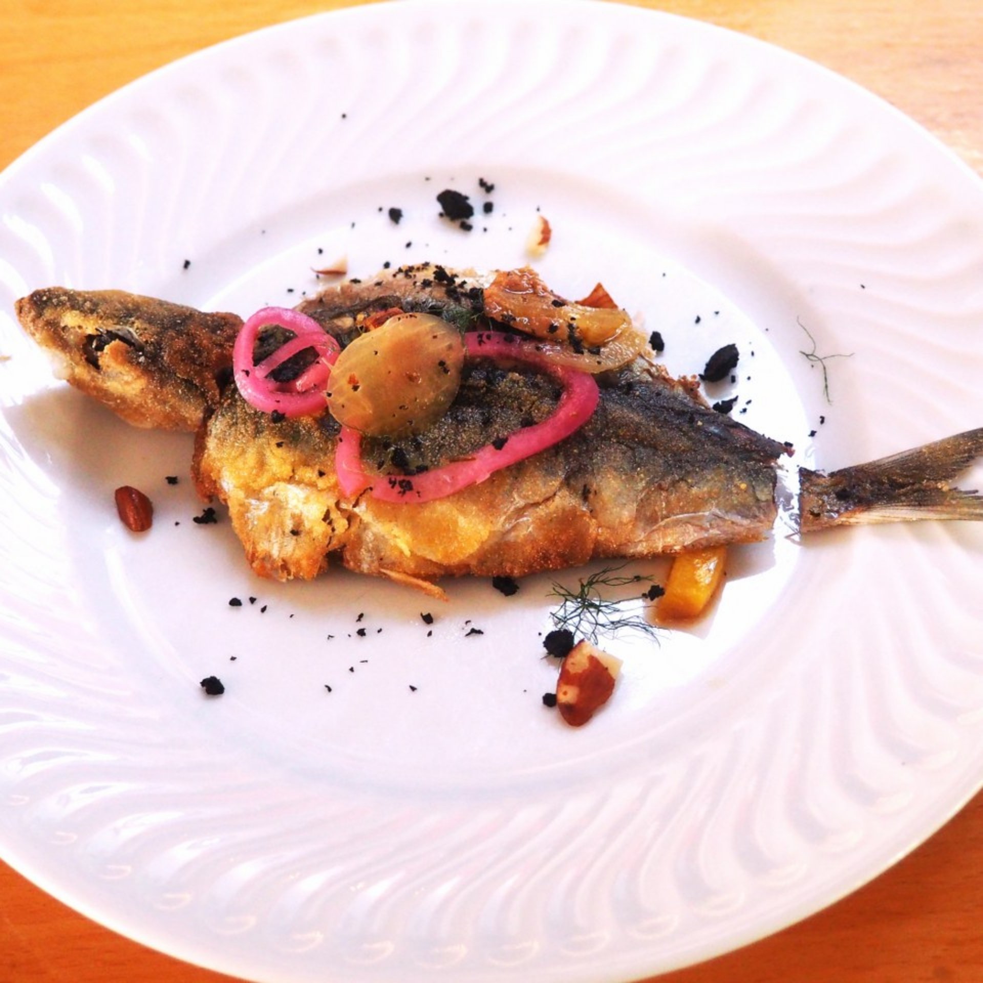 Pescado frito entero en un plato blanco, aderezado con cebolla roja encurtida, aceitunas verdes y migas de especias negras