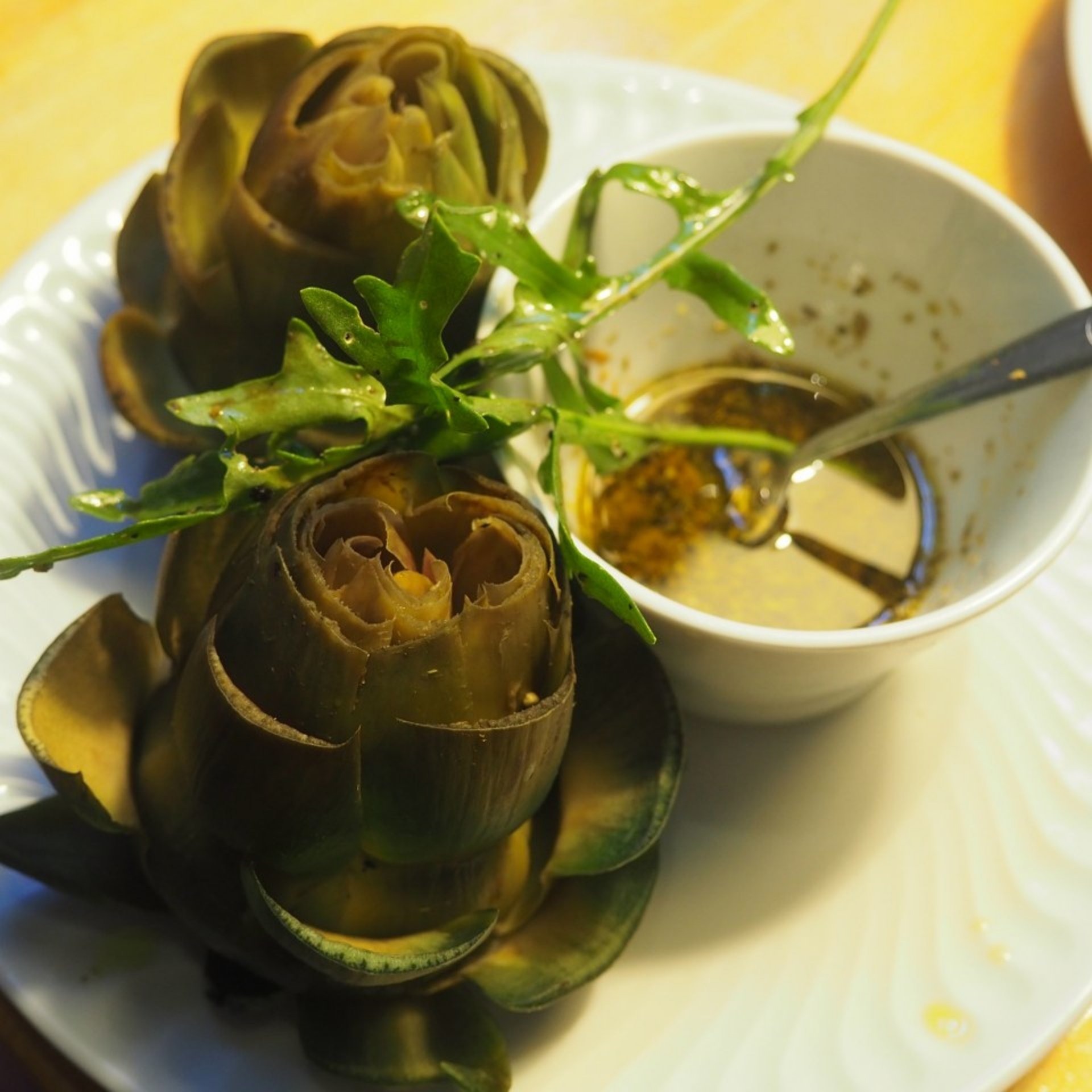 Dos alcachofas al vapor adornadas con rúcula en un plato blanco, acompañadas de una salsa para mojar en un cuenco pequeño