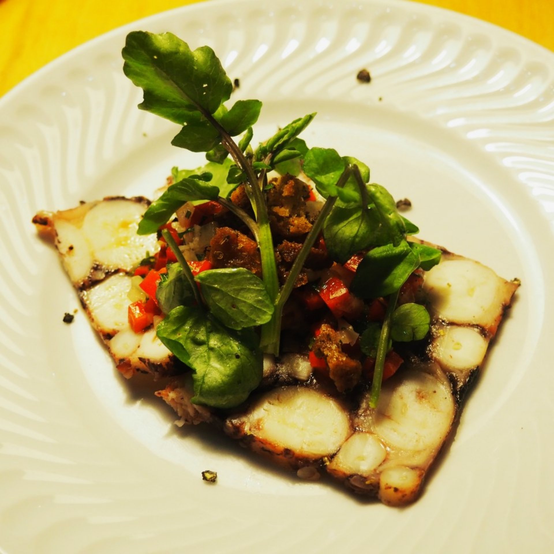 Pulpo a la parrilla en un plato blanco, aderezado con pimiento rojo cortado en dados, pan rallado y hojas frescas de berro