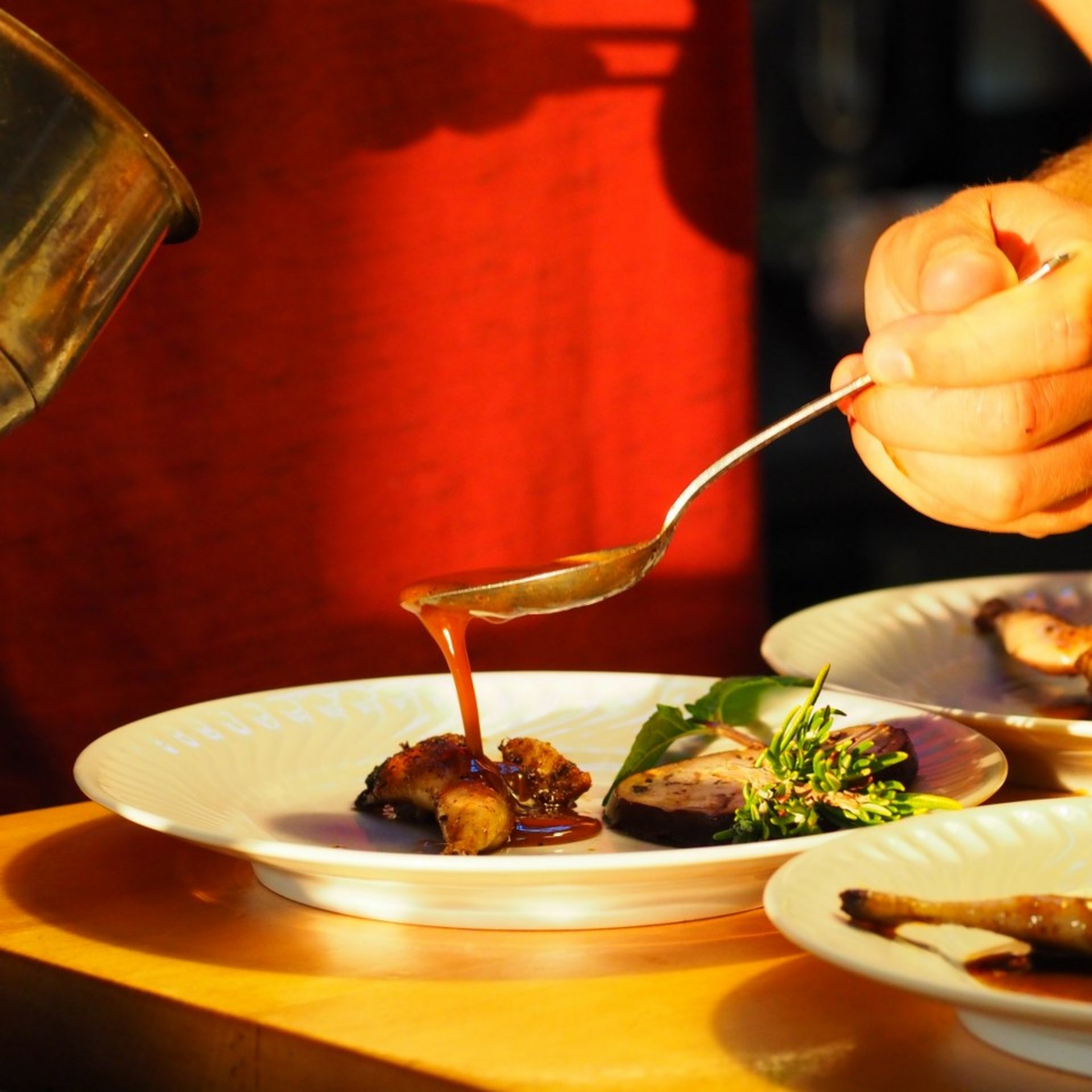 En un restaurante acogedor y con luz tenue, un chef vierte con un cucharón una salsa espesa y sabrosa sobre un plato de carne y hierbas frescas.