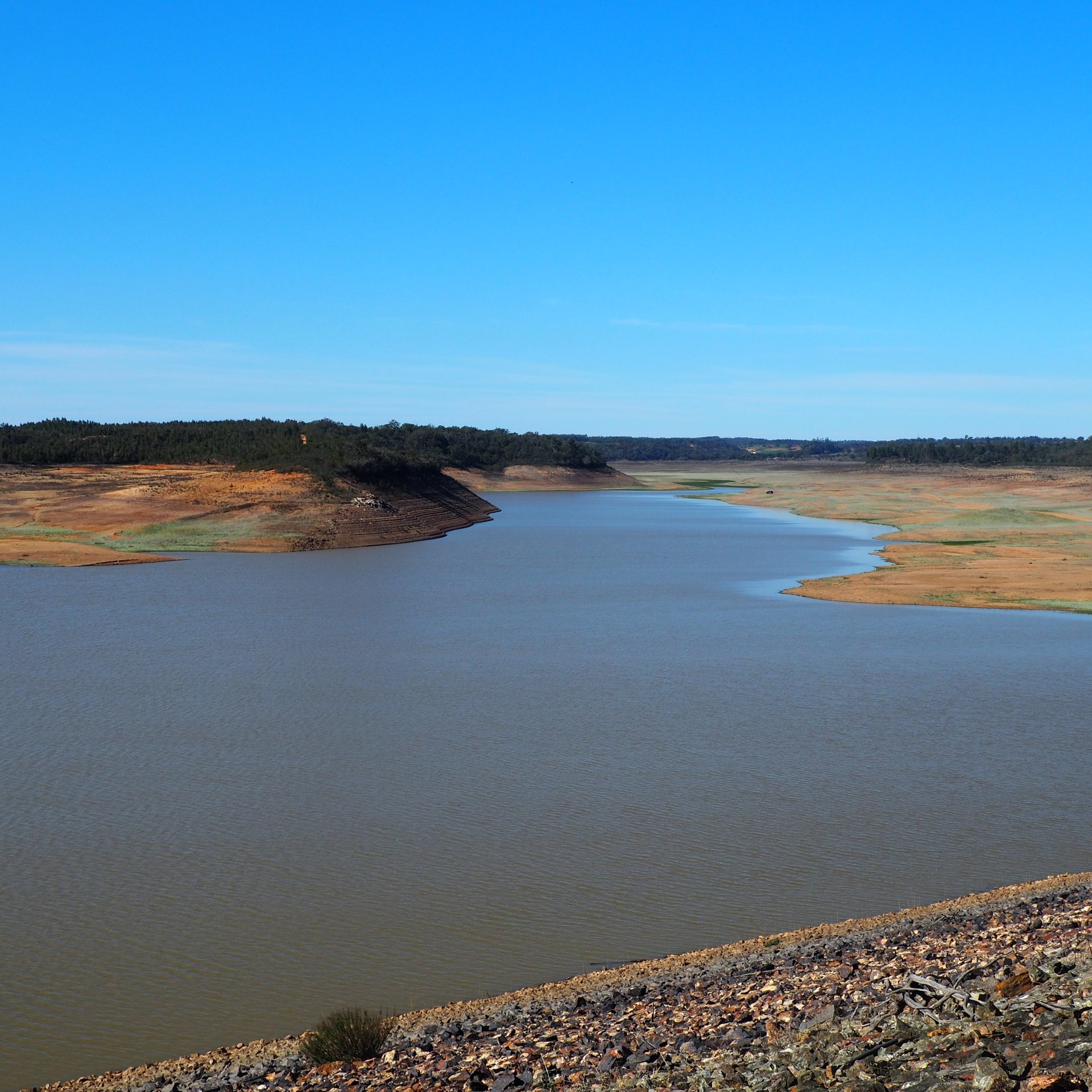 Embalse con niveles de agua críticamente bajos, con la orilla seca y árida al descubierto y el terraplén rocoso de la presa visible en primer plano