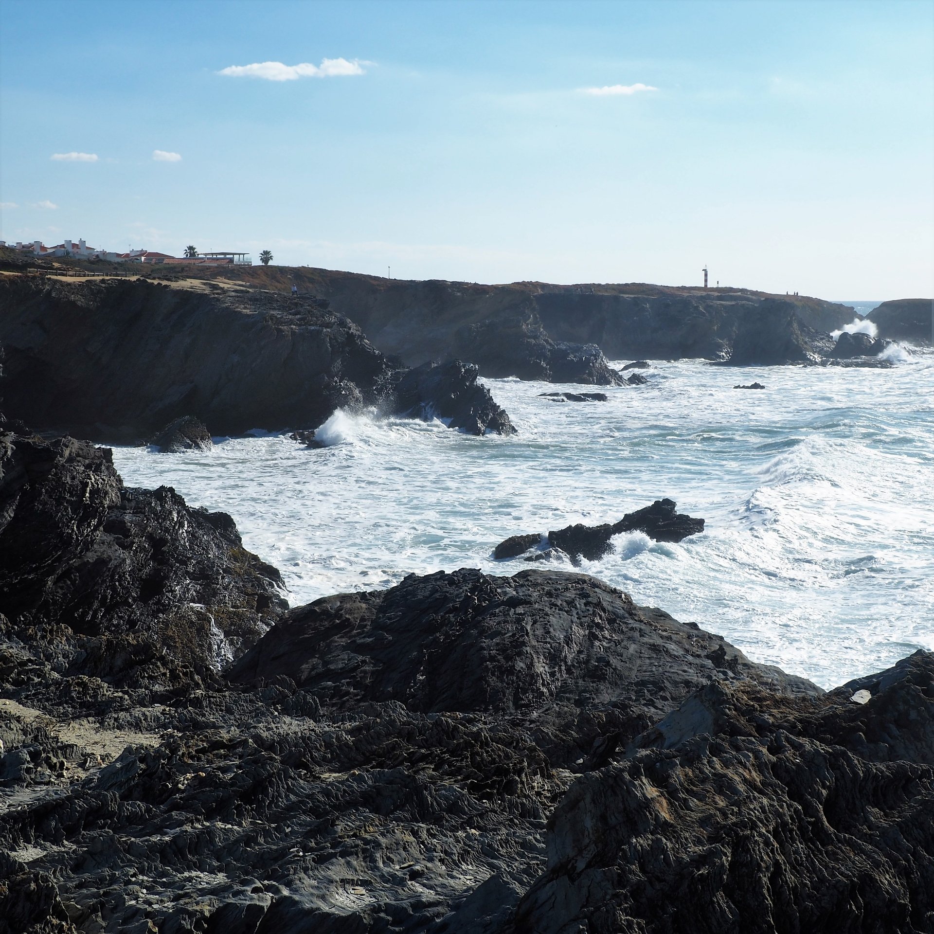 Una costa rocosa con olas rompiendo, un acantilado con edificios y un faro visible al fondo bajo un cielo azul