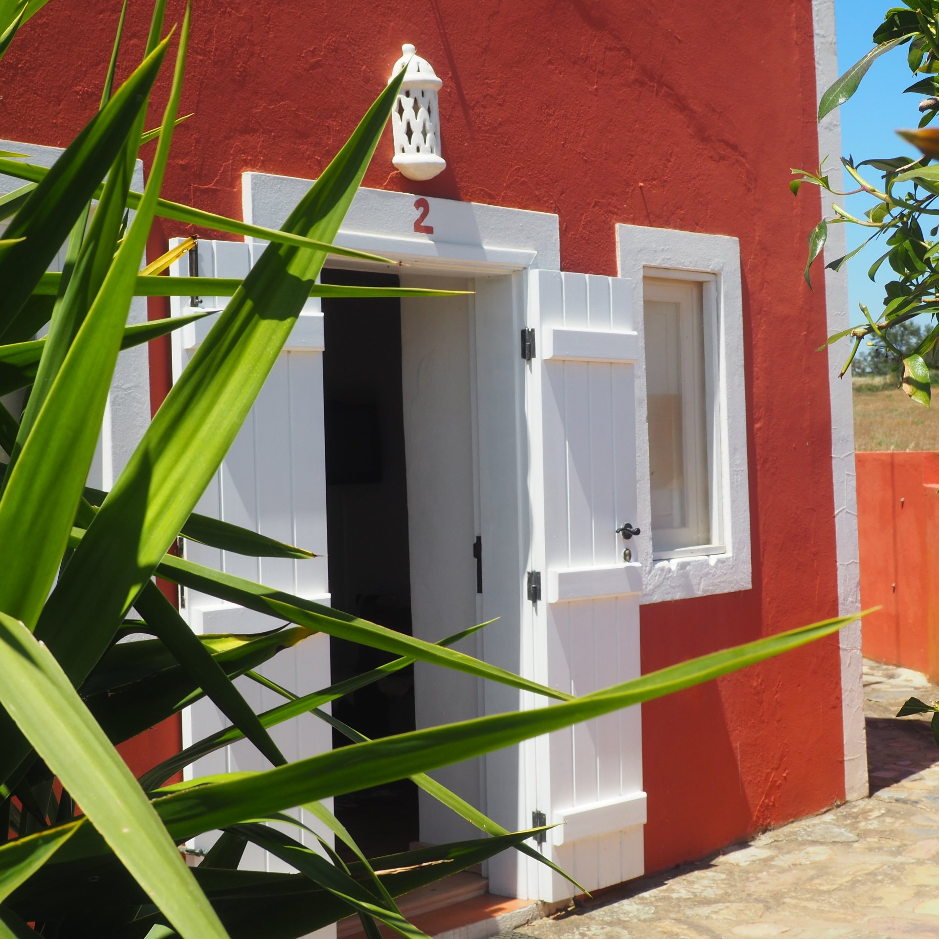 Exterior de la vivienda número 2, un edificio pintado de rojo con una puerta holandesa blanca y una linterna de pared, enmarcado por hojas de palmera