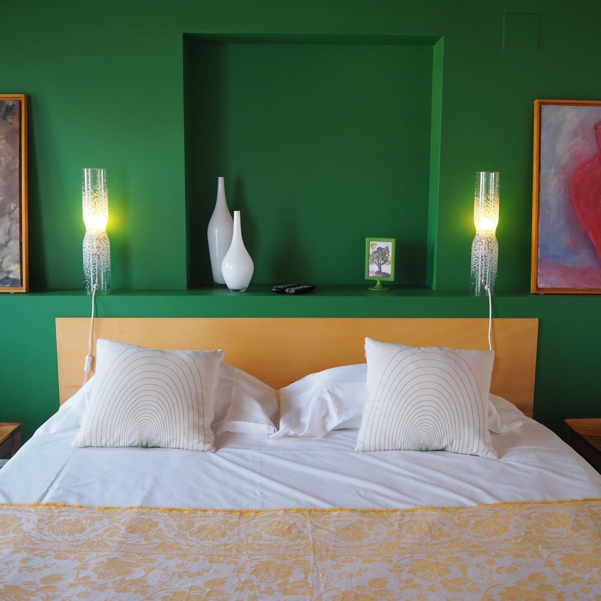 Habitación de hotel con cama de matrimonio, paredes de un verde intenso, un nicho empotrado en la pared con jarrones blancos y lámparas de pared cilíndricas que emiten una luz cálida a ambos lados