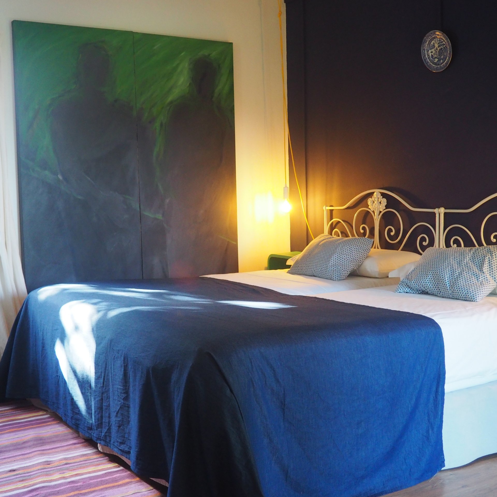 Habitación de hotel con una cama de matrimonio, cabecero de hierro blanco, colcha azul y un gran cuadro abstracto verde en la pared