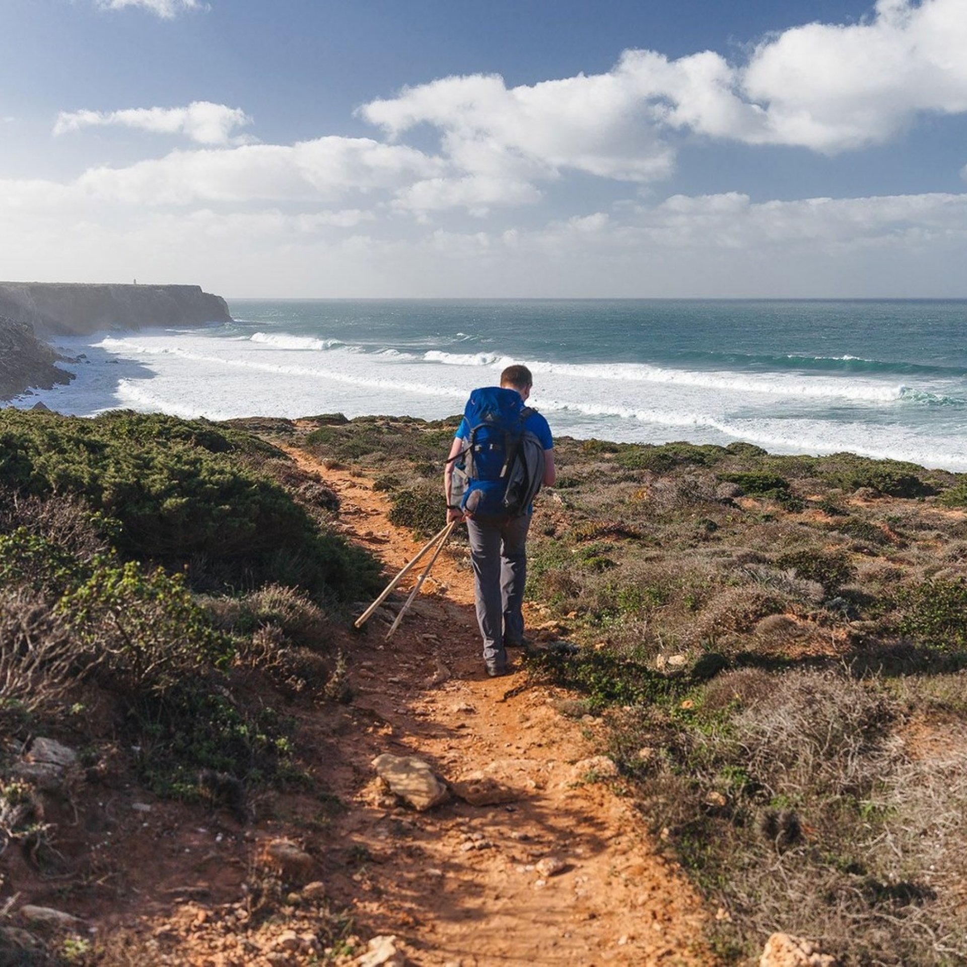Excursionista con una mochila grande y bastones de senderismo que recorre un sendero costero de tierra, con las olas rompiendo y los acantilados rocosos a sus pies