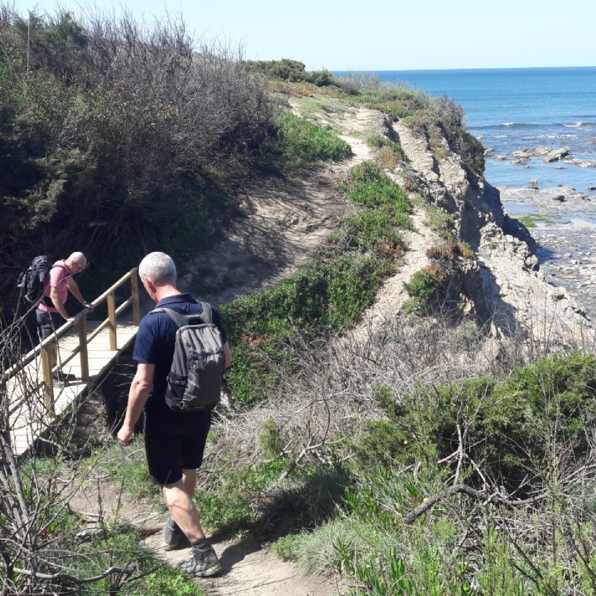 Dos excursionistas con mochilas cruzando un puente peatonal de madera en un sendero costero sobre un acantilado, con la costa rocosa y el océano azul a sus pies