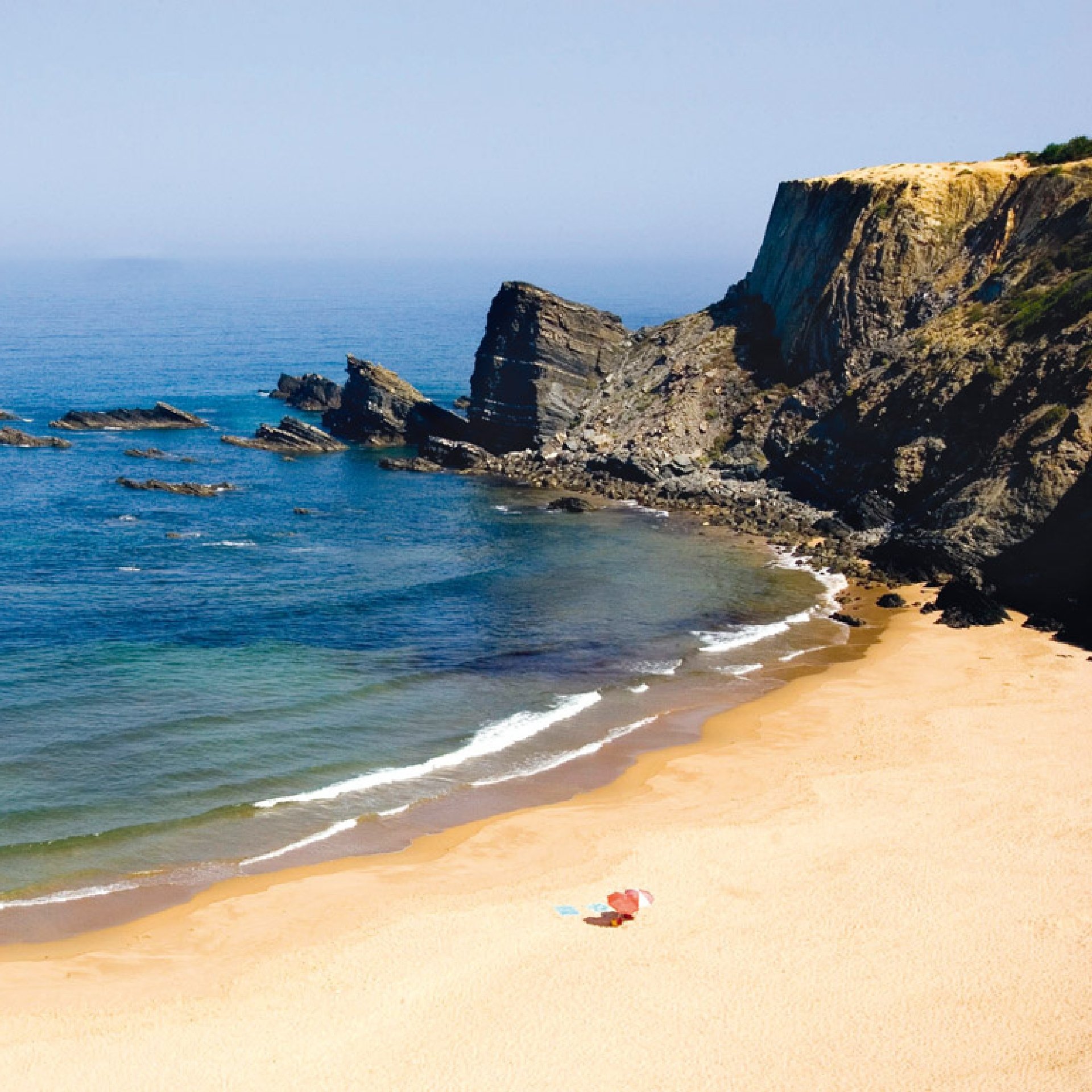 Una cala de arena apartada con espectaculares acantilados oscuros, afloramientos rocosos en el océano azul y una toalla de playa solitaria en la orilla