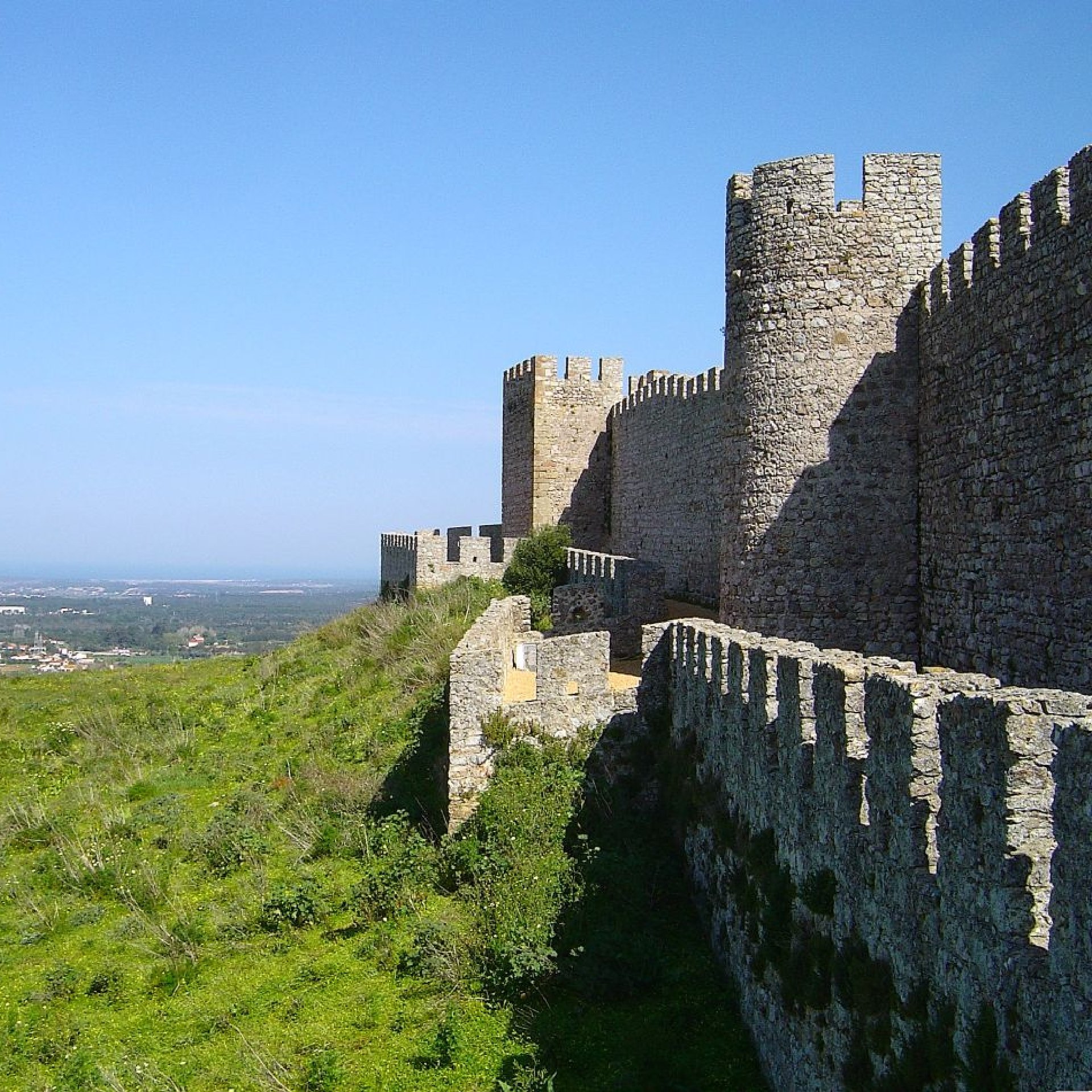 Murallas de un castillo medieval y torres almenadas en lo alto de una colina, con una ladera verde y un paisaje costero en la lejanía a sus pies