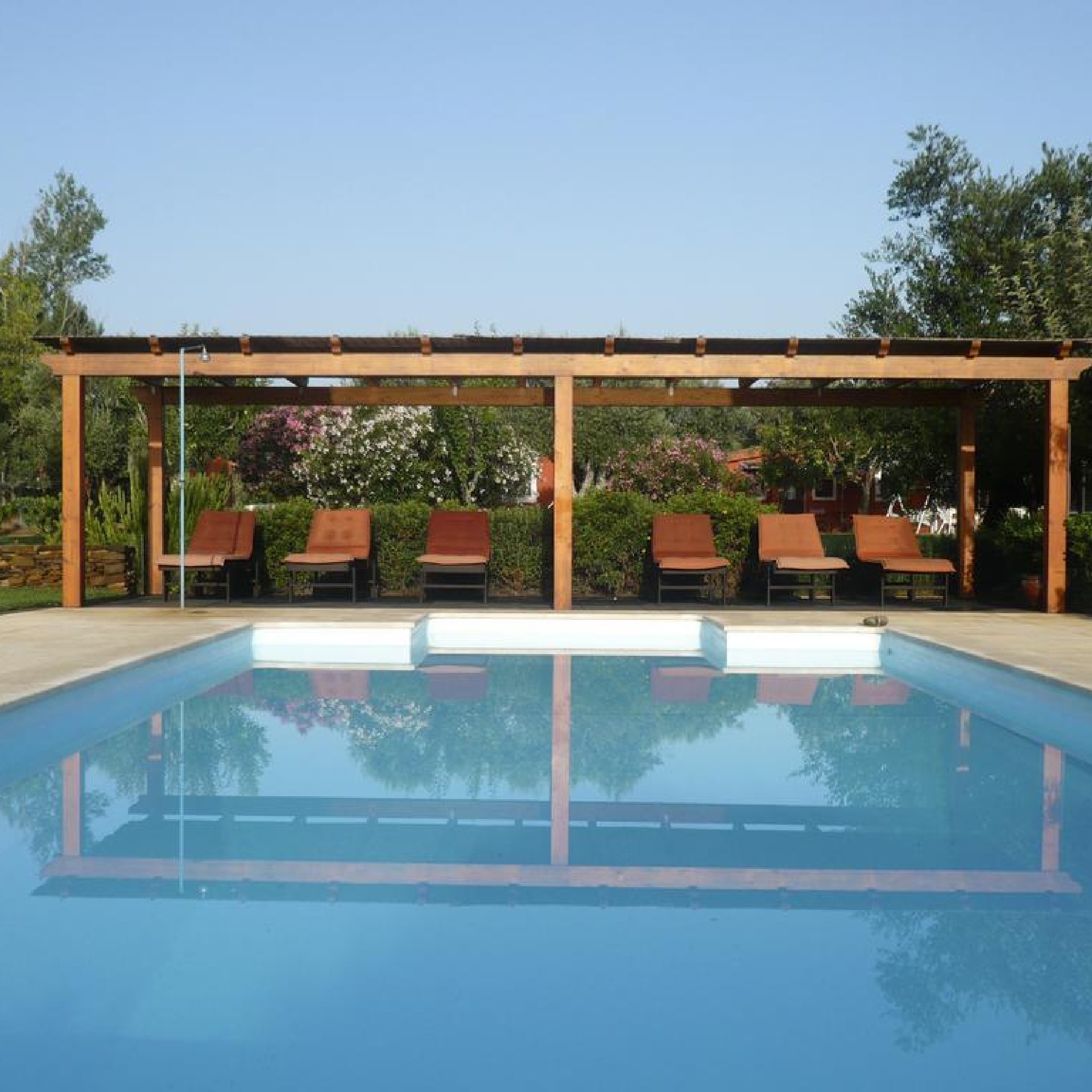 Piscina al aire libre con tumbonas bajo una pérgola de madera, rodeada de exuberante vegetación y arbustos en flor