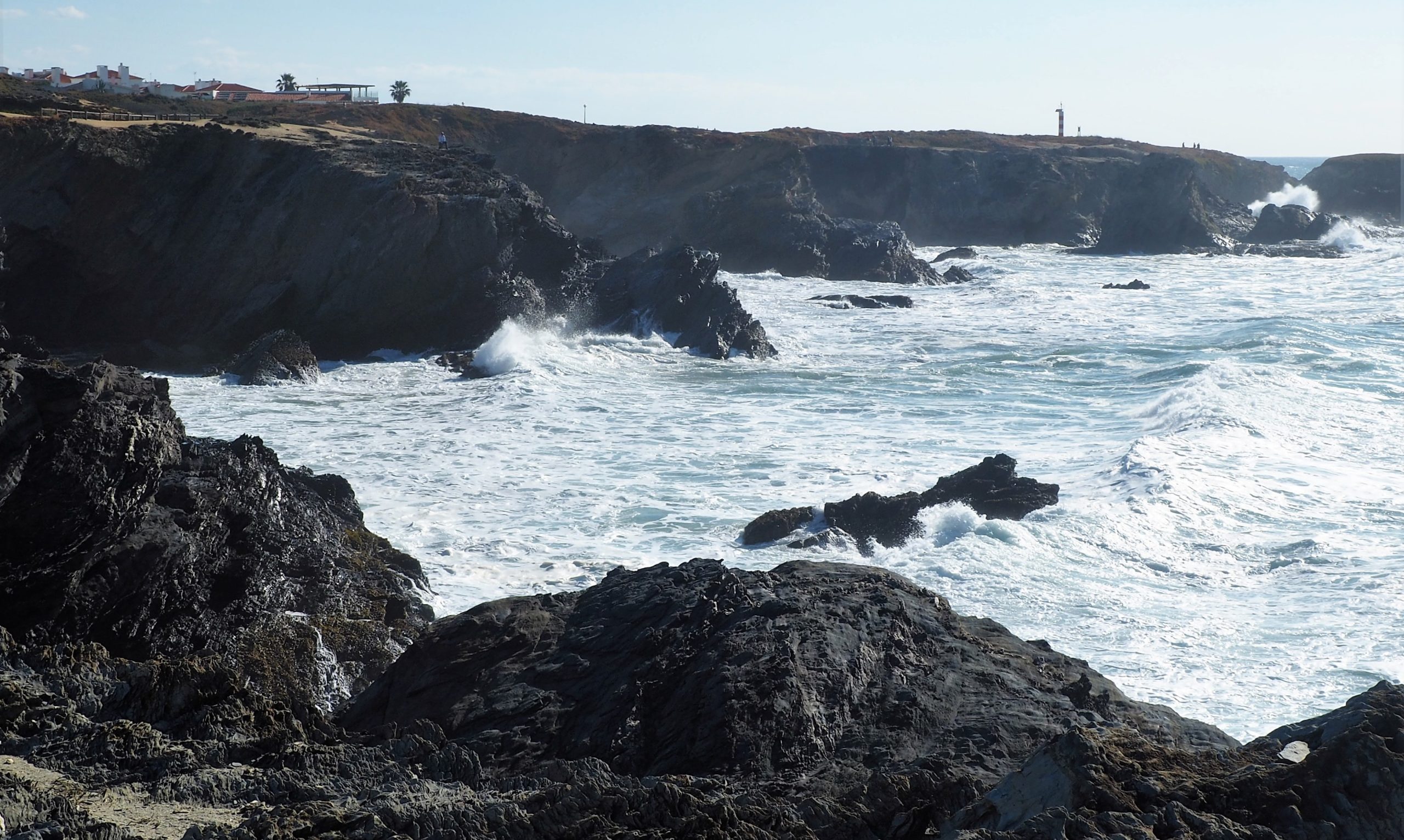 Una costa rocosa con olas rompiendo contra las rocas oscuras; en primer plano, un acantilado con edificios y un faro visible al fondo