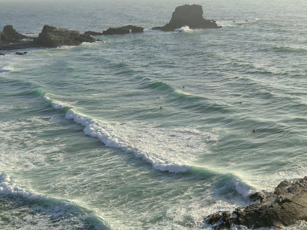 Surfer im Meer mit rollenden Wellen und felsigen Ausläufern entlang einer zerklüfteten Küste