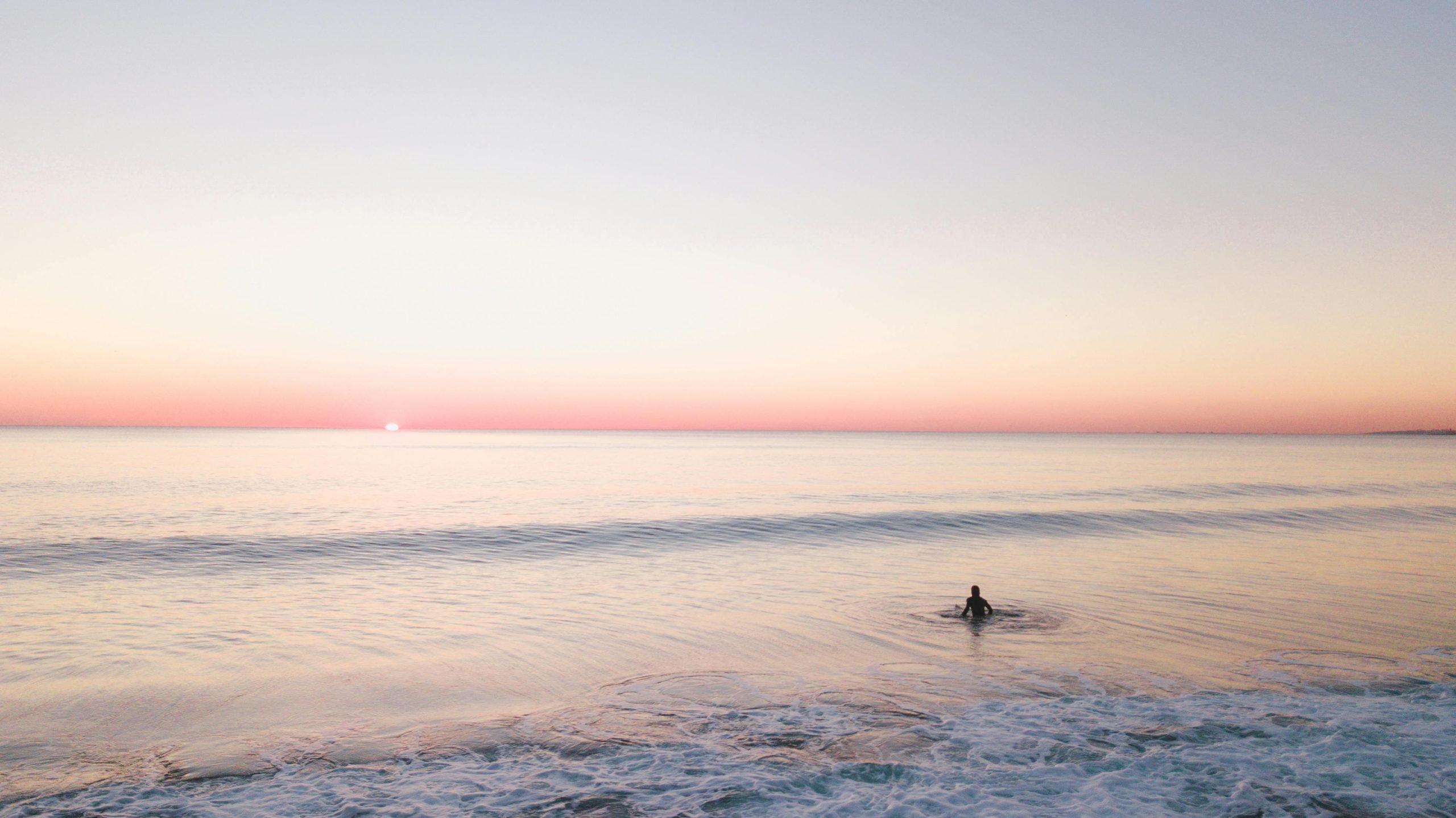 Eine einsame Person, die bei Sonnenuntergang im ruhigen Wasser des Ozeans watet, mit einem pastellrosa und orangefarbenen Horizont, der sich über den Himmel erstreckt