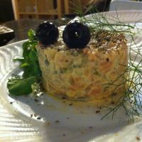 Tártaro de salmão moldado, coberto com duas azeitonas pretas e endro fresco, servido com verduras num prato branco