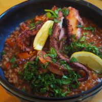 Polvo grelhado servido com arroz de tomate numa tigela escura, guarnecido com coentros frescos e rodelas de limão